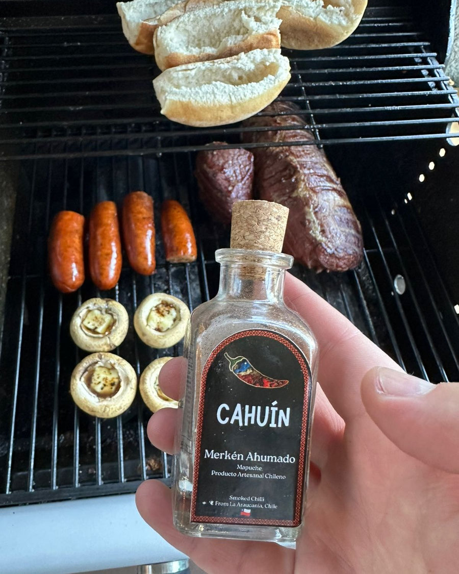 Botella de MERKÉN CAHUÍN en la mano frente a una parrilla con choripanes, carne y champiñones